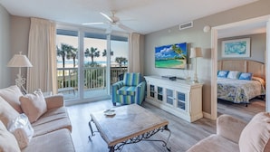 A spacious living room with a large sliding glass door to a balcony, a ceiling fan, a flat-screen TV, a sofa, a coffee table, and a bed with a view of the beach. - Pelican Beach 0216 2 Bedroom Condo by Pelican Beach Management (Destin)