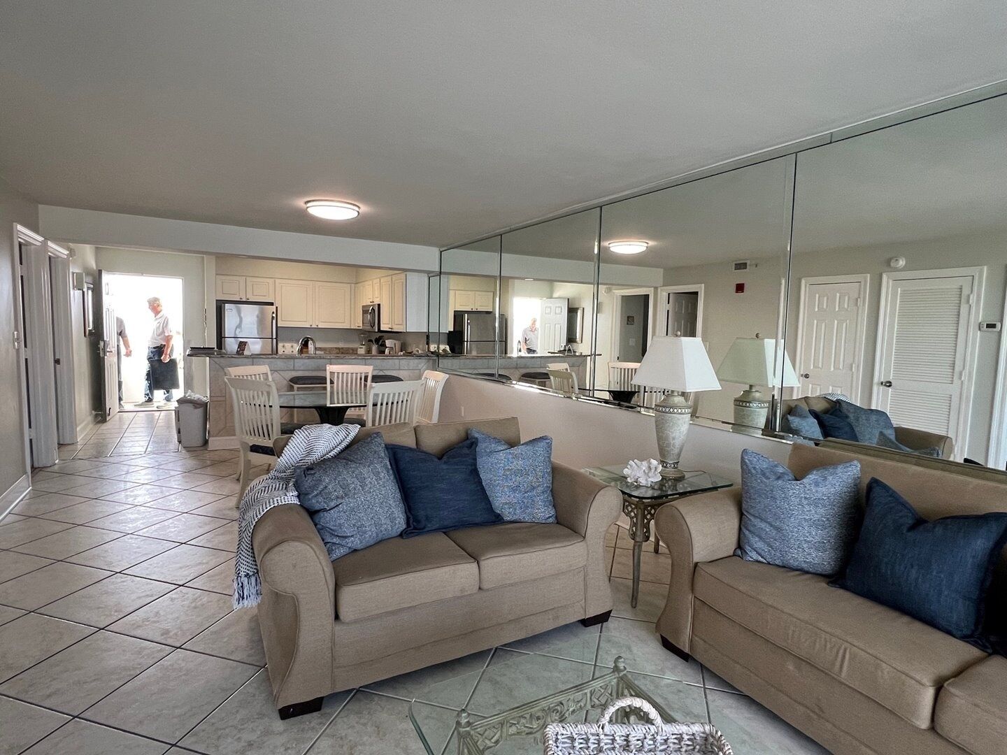 A living room with a beige sofa, a glass coffee table, and a large mirror reflecting the kitchen area.