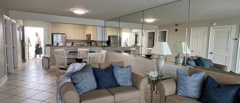 A living room with a beige sofa, a glass coffee table, and a large mirror reflecting the kitchen area.