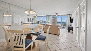 A spacious living area with a large glass door leading to a balcony, a dining area with a glass table, and a ceiling fan. - Pelican Beach 1414 2 Bedroom Condo by Pelican Beach Management (Destin)