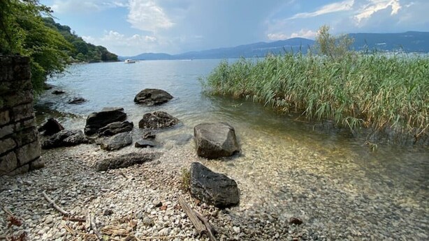 Un rivage rocheux bordé d’une eau claire, un bateau au loin et une vue sur des montagnes en arrière-plan.