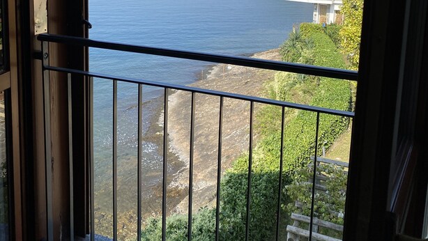 A balcony with a view of a calm lake, a rocky shoreline, and a hill with buildings. - Gemütliche 3 Zimmer Fewo mit Traumblick (Leggiuno)