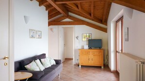 A cozy living room with a wooden ceiling, a sofa with floral cushions, a TV on a wooden cabinet, and a radiator on the wall. - Gemütliche 3 Zimmer Fewo mit Traumblick (Leggiuno)