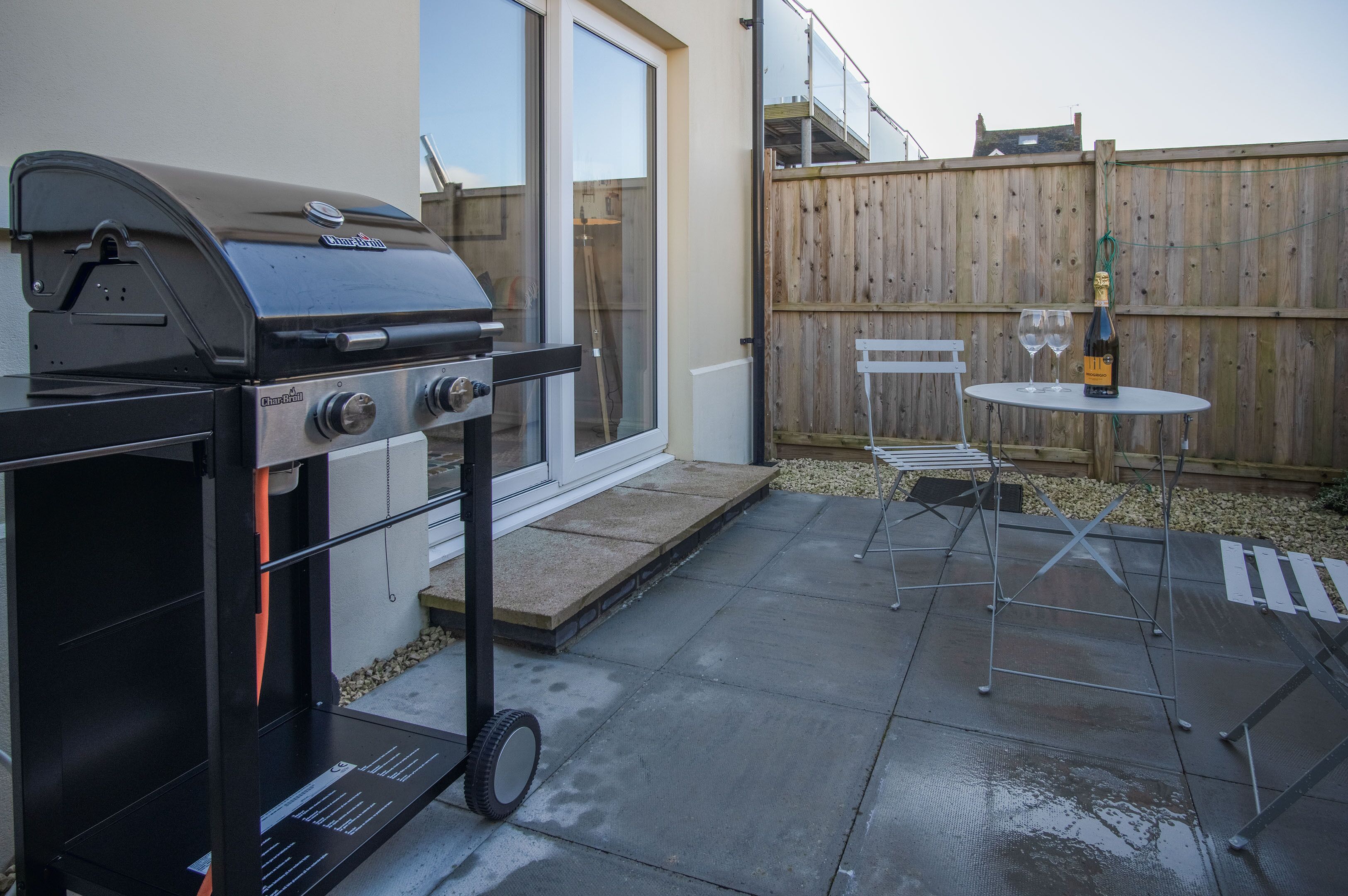 A backyard with a barbecue grill, a table with two glasses, and a bottle of wine.