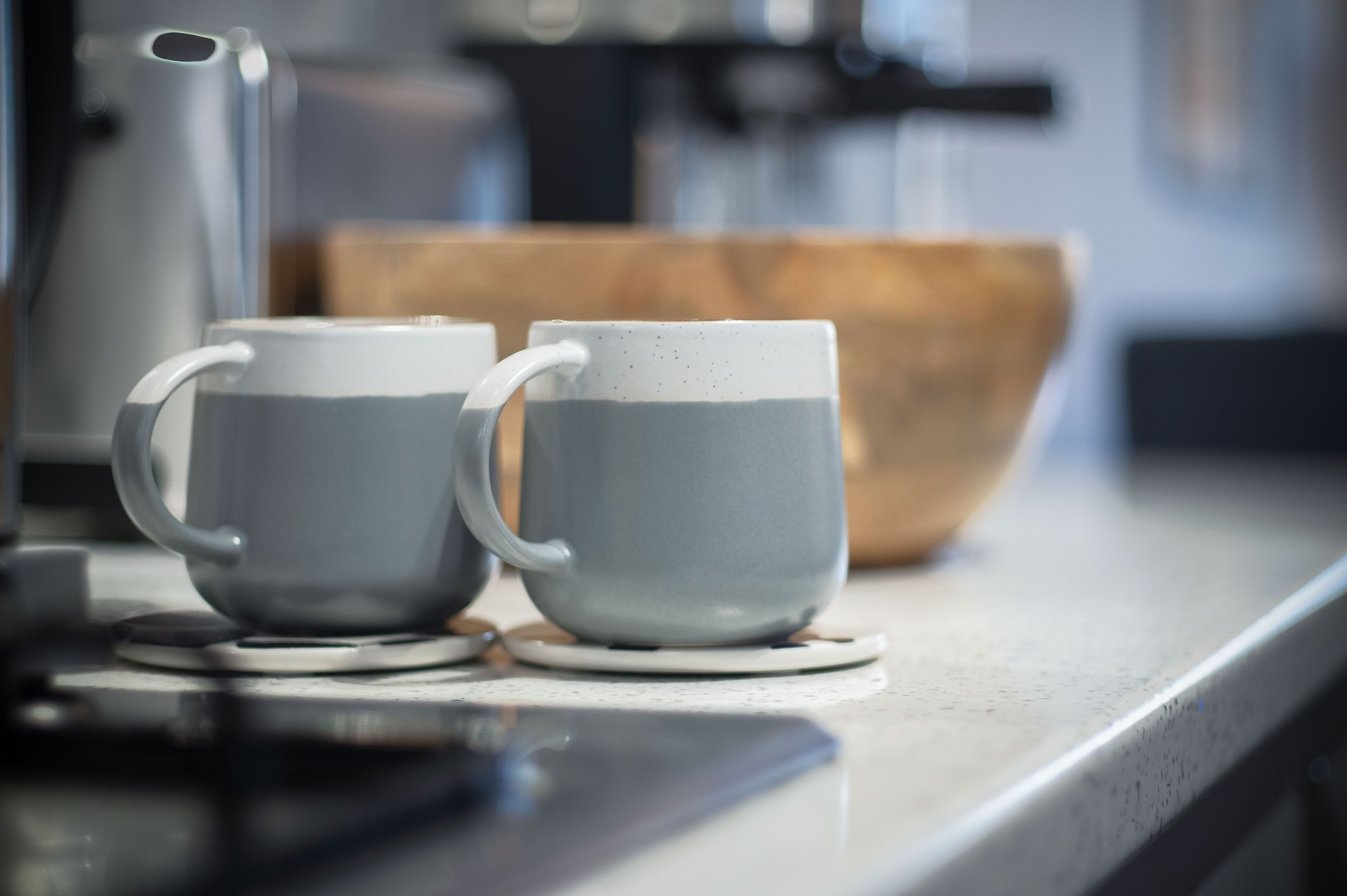 Twee grijze keramische bekers op een witte onderzetter, een houten schaal en een roestvrijstalen waterkoker op een keukentafel.
