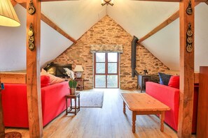 Ein gemütliches Dachzimmer mit Steinwand, Holzbalken, einem roten Sofa, einem Holztisch und einem Fenster mit Blick.