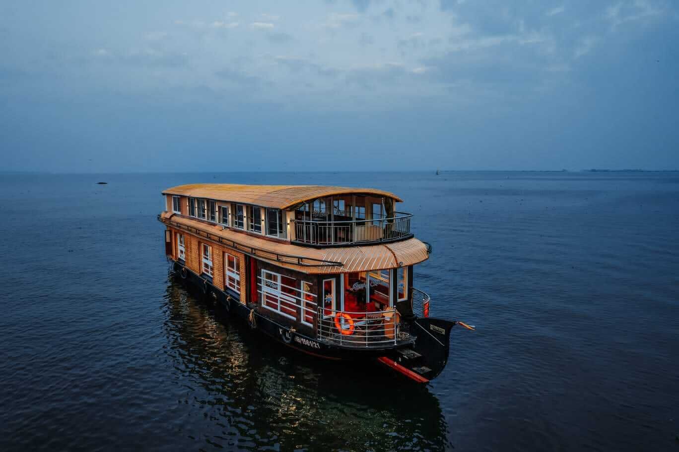 A large, multi-deck houseboat with a wooden deck and multiple windows, floating on calm water.