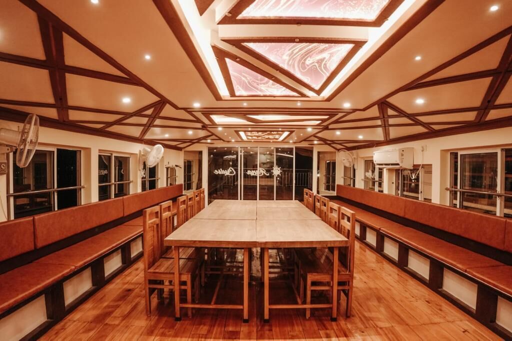 A spacious dining area with a long wooden table and benches, large windows, and ceiling fans.