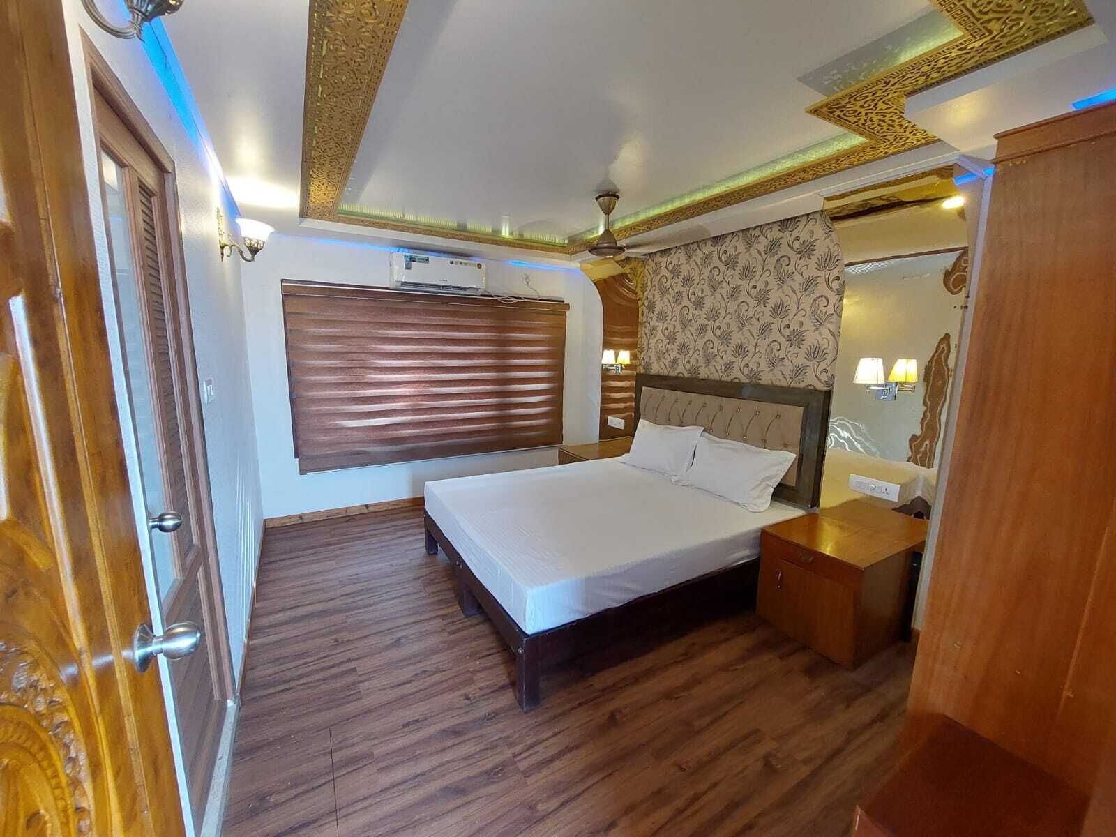 A bedroom with a wooden floor, a bed with a headboard, a window with blinds, a wall-mounted air conditioner, and a nightstand with a lamp.