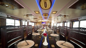 Vagón comedor de tren con mesas preparadas para comer, un pasillo central y una pantalla grande al frente.