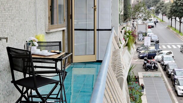 A balcony with a black chair, a small table with a white cup and a plate of food, and a view of a street with parked cars and buildings. - 4bnb - Cozy Albornoz Flat (Rome)