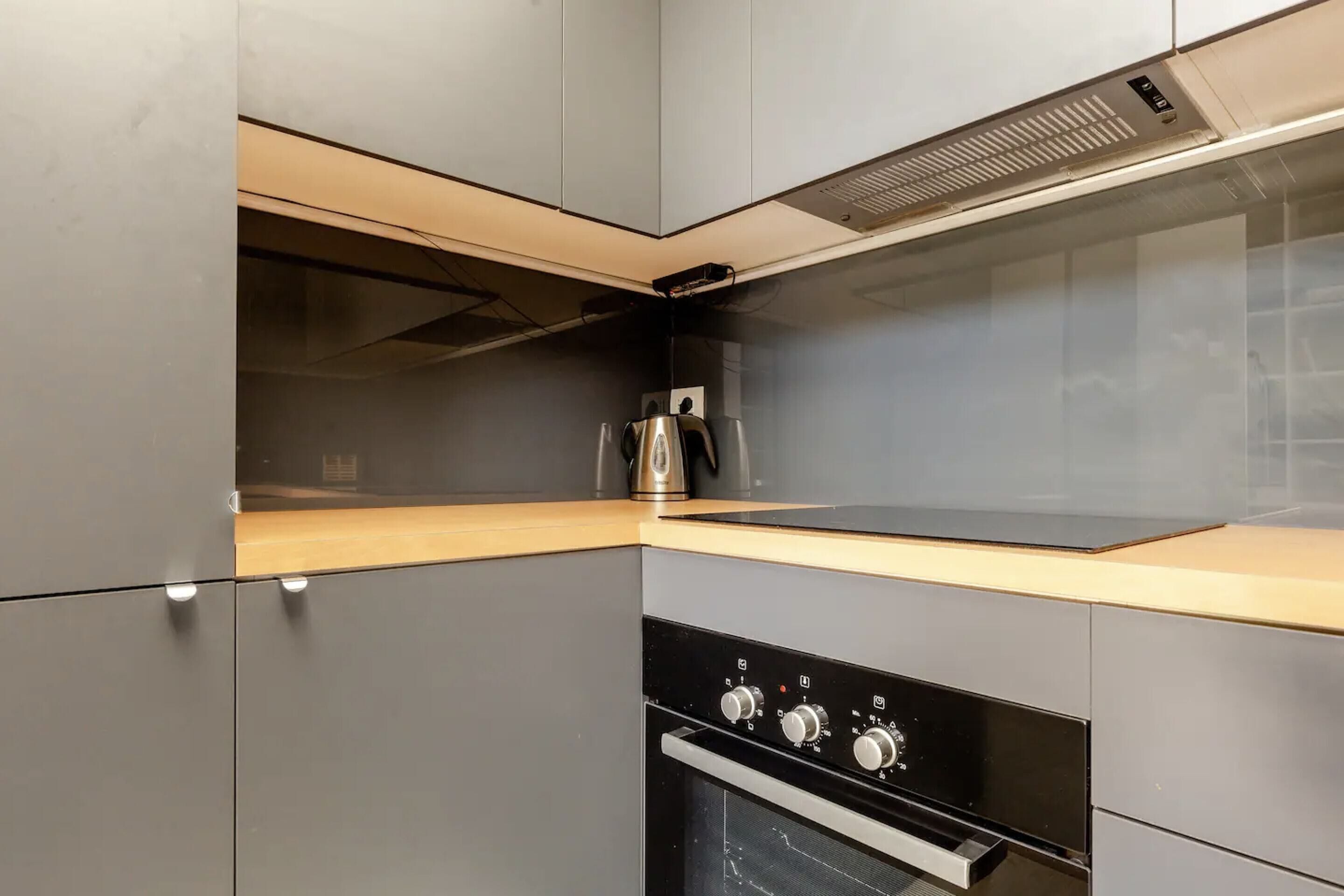 A modern kitchen with grey cabinets, a black oven, and a stainless steel kettle on the countertop.
