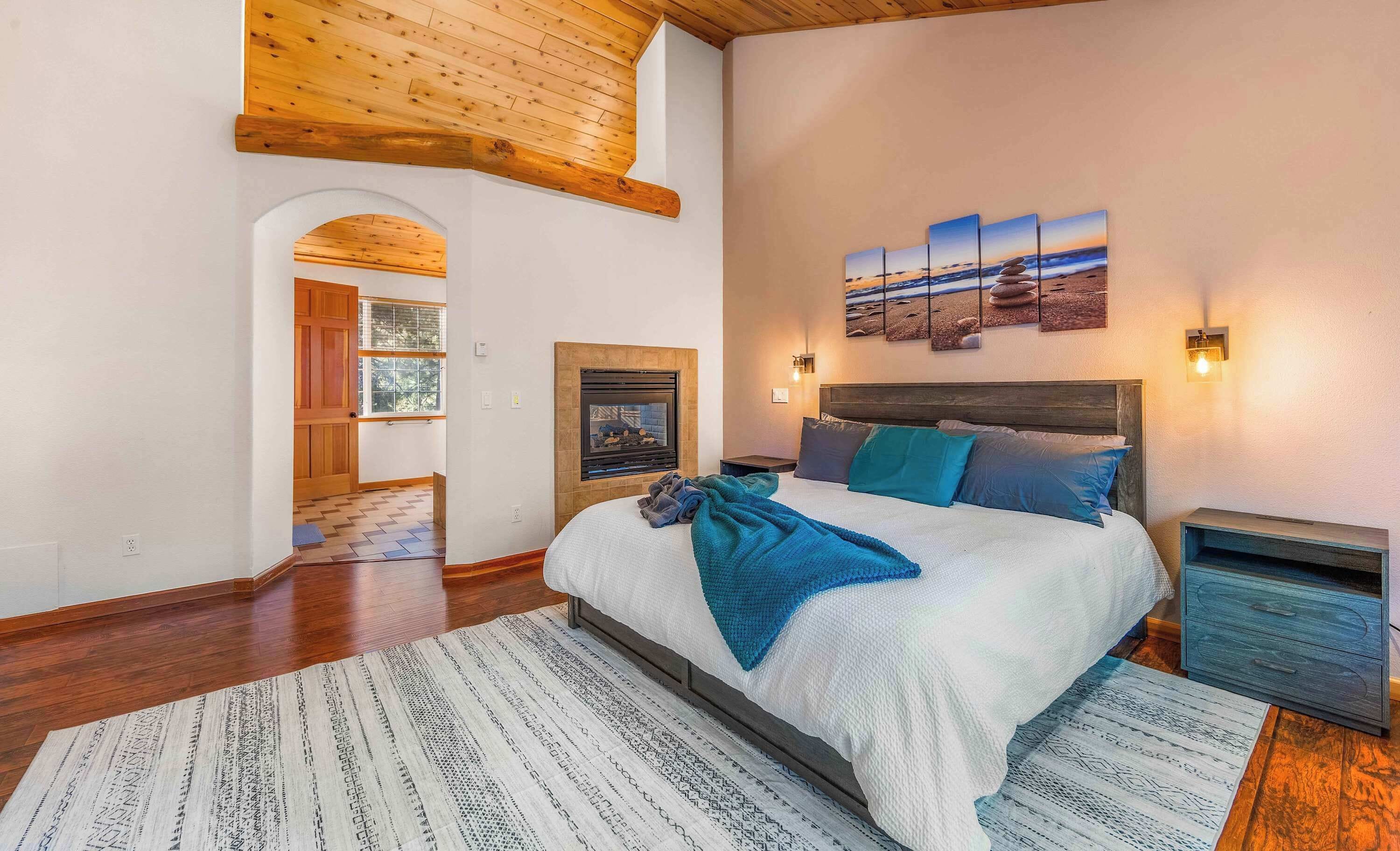 A bedroom with a large bed, a fireplace, a wooden ceiling, and a view of the outdoors.
