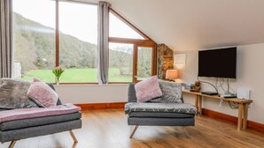 A room with two grey armchairs, a wooden floor, a TV on a wooden stand, and a window with a view of greenery. - Monkleigh Mill (Bideford)