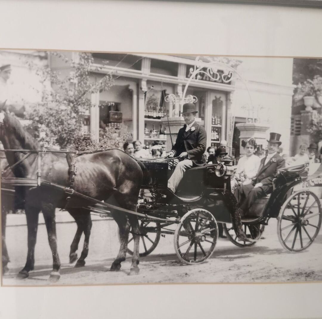 Een oude foto van een koets getrokken door paarden, met passagiers, waaronder een man met een hoge hoed en een vrouw met een hoed, voor een gebouw met een winkel en een bord.