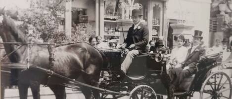 A vintage photograph of a horse-drawn carriage with passengers, including a man in a top hat and a woman in a bonnet, in front of a building with a shop and a signboard.