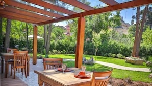 Un patio exterior con mesas y sillas de madera, una pérgola en lo alto y vistas de un jardín ajardinado.