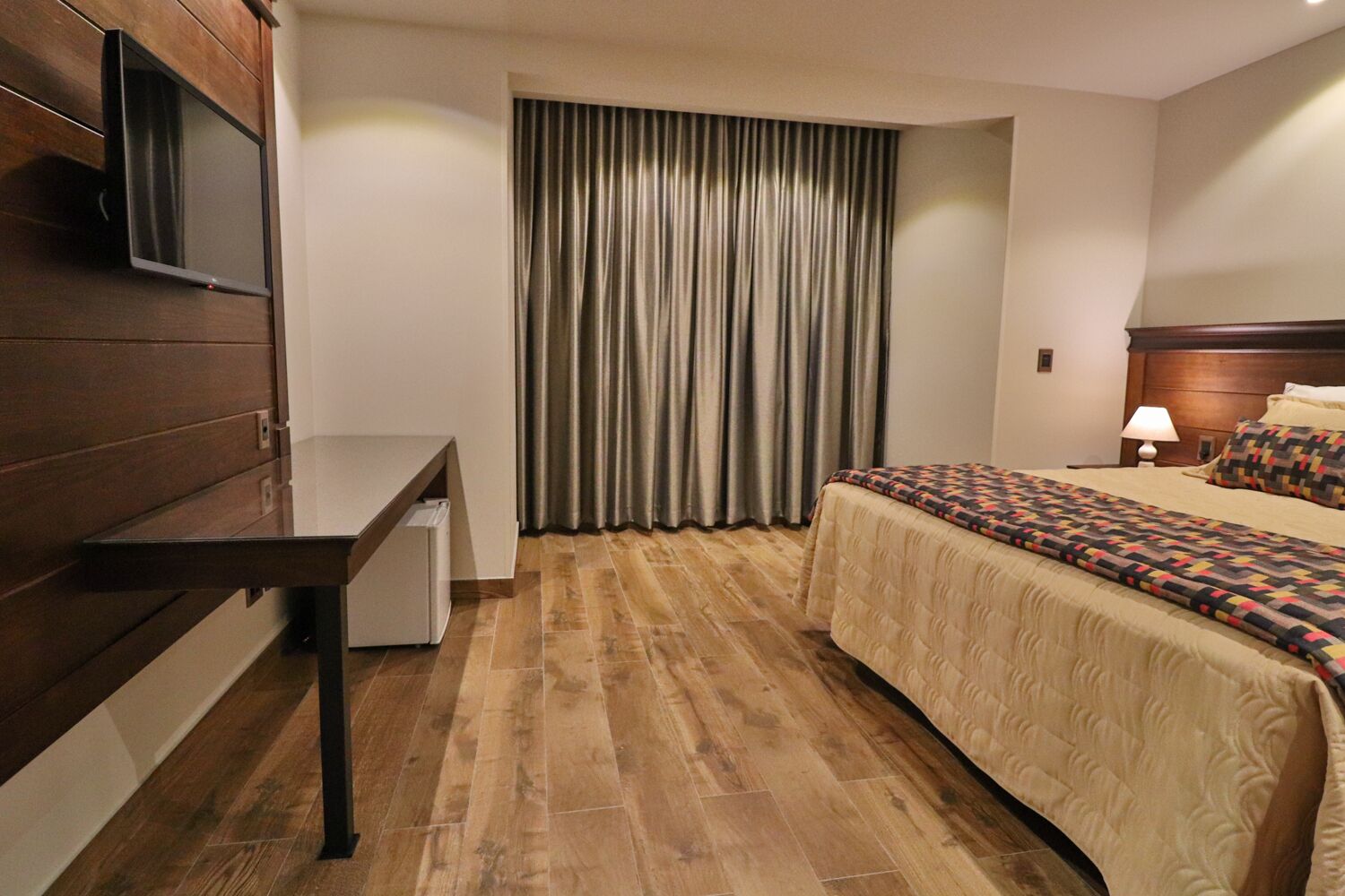 Una habitación de hotel con cama, escritorio, televisión y ventana con cortinas.