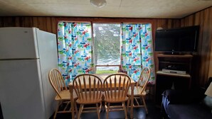 Eine kleine Küche mit einem Kühlschrank, einem Holzespeisetisch mit Stühlen, einem Fernseher auf einem Schrank und einem Fenster mit blumigen Vorhängen.