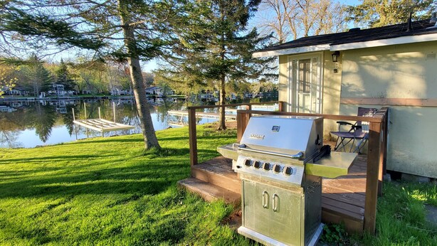 Ein tragbarer Grill auf einer Holzterrasse an einem Seeufer, mit einem kleinen Schuppen und einem Steg im Hintergrund.