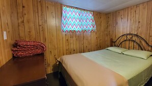 Ein Zimmer mit Holzwänden, einem Bett mit grüner Bettdecke, einem Fenster mit bunter Zickzack-Gardine und einem Spiegel an der Wand.