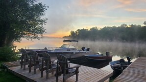 Eine Holzsteg mit Stühlen und einem Boot, umgeben von Bäumen und einem nebligen See im Morgengrauen.