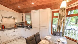 A modern kitchen with white cabinets, a wooden ceiling, and a dining area with a table set for four. - Cedar Lodge (Windermere)