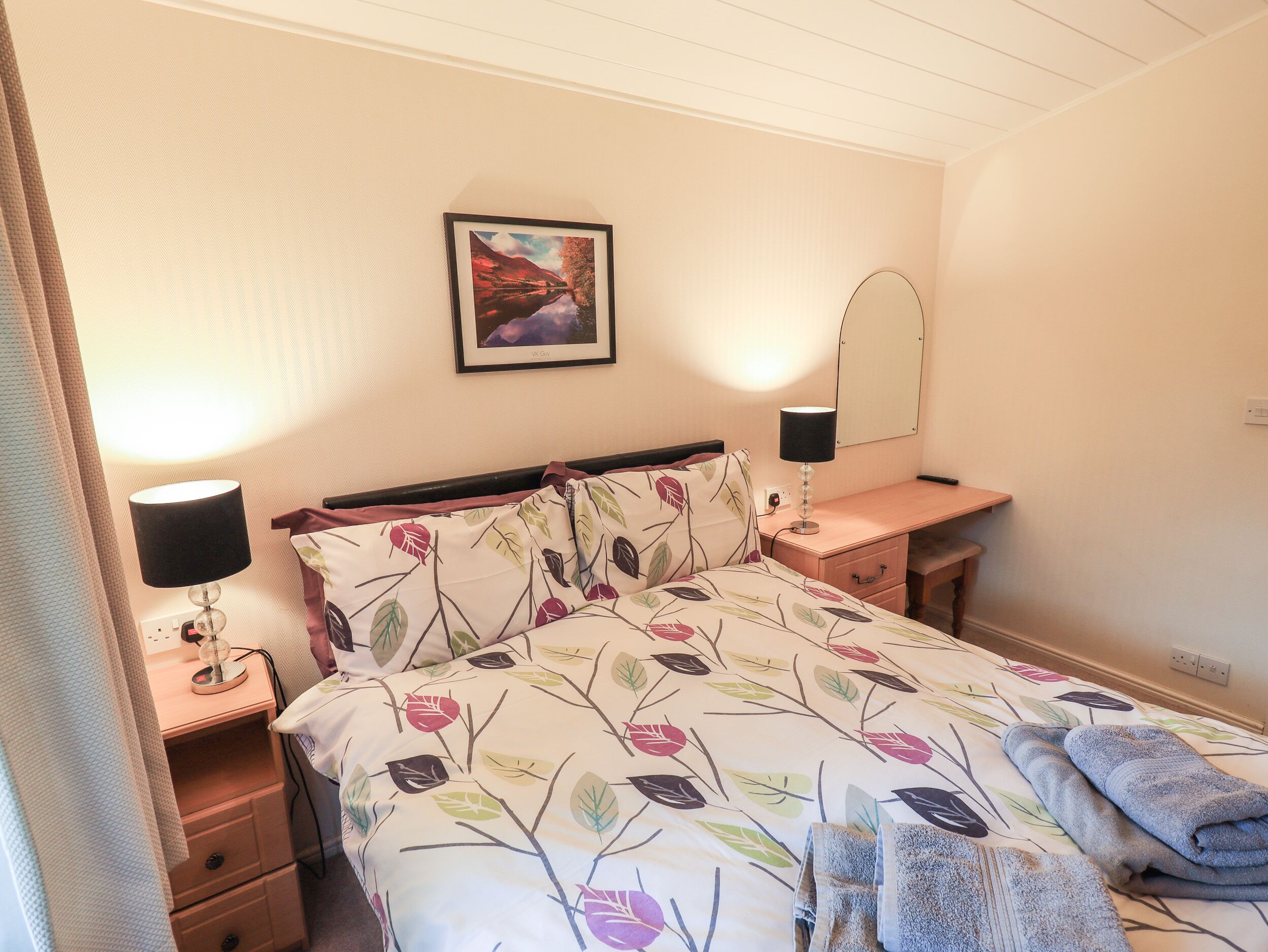 A neatly arranged bedroom with a bed, a bedside table with a lamp, a mirror, and a wall-mounted picture.