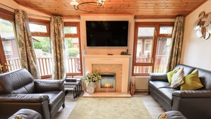 A living room with a fireplace, leather sofas, a flat-screen TV, and large windows with curtains. - Cedar Lodge (Windermere)