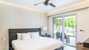 Habitación de hotel moderna con una cama grande, ventilador de techo, mesita de noche, lámpara y vistas al exterior a través de una puerta corrediza de vidrio.