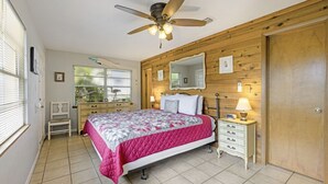 Ein Schlafzimmer mit Bett, Nachttisch, Schminktisch, Deckenventilator, Fenster und Holzvertäfelung an den Wänden.