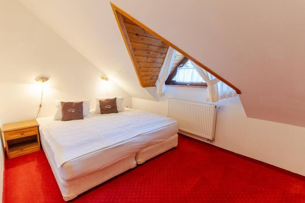 Ein Schlafzimmer mit schrägen Wänden, einem Bett mit weißer Bettwäsche, zwei Kissen, einem Nachttisch, einem Heizkörper und einem Fenster mit Vorhängen.