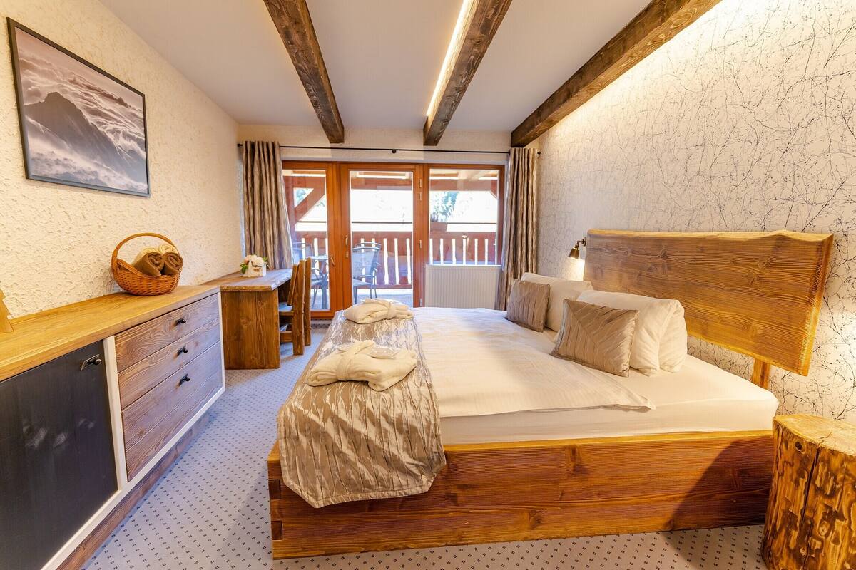 Ein Schlafzimmer mit einem hölzernen Bett, einem Nachttisch, einer Bank, einer Holzkommode, einem Fenster mit Vorhängen und Blick auf den Außenbalkon.