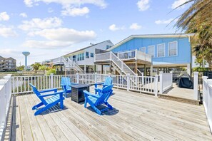 A wooden deck with blue Adirondack chairs and a table, a blue house with a white fence, and a water tower in the background. - Charlie's Beach Retreat | Fun 4BR w/ Heated Pool (North Myrtle Beach)