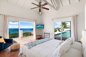 A bedroom with a bed, a ceiling fan, and a TV. There are two large windows with views of the ocean and a pool.