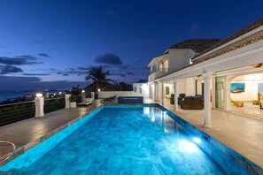 A swimming pool with a view of the ocean, a covered patio area, and a building with a balcony.