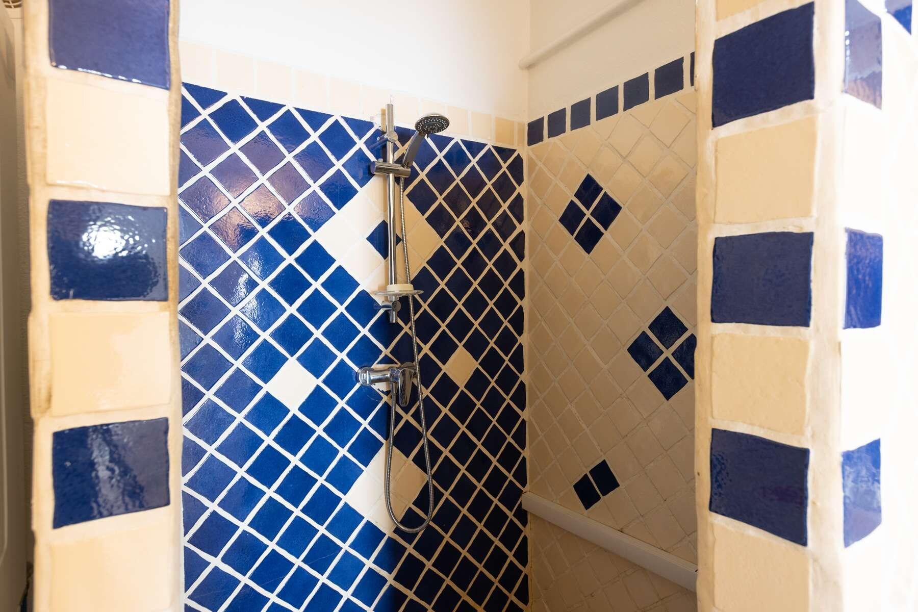 Un baño con baldosas azules y beige estampadas, una ducha con cabezal de ducha manual y una bañera blanca.