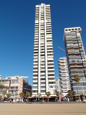 Een hoog, modern hoogbouwgebouw met meerdere balkons en een helderblauwe lucht op de achtergrond.