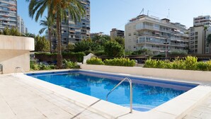 Una piscina amb terrassa enrajolada, envoltada de vegetació i palmeres, amb edificis alts al fons.