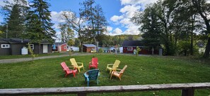 A grassy area with colorful Adirondack chairs arranged in a circle, surrounded by wooden cabins and a lake in the background. - River View 3 Bedroom Cottage 12 (Hastings)