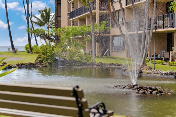 Un élément décoratif aquatique comprenant une fontaine, situé dans un espace paysager agrémenté d’un banc, de palmiers et d’un bâtiment doté de balcons.
