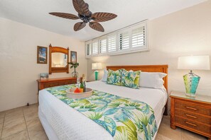 A bedroom with a bed, a ceiling fan, a nightstand with a lamp, a dresser, and a window with shutters.