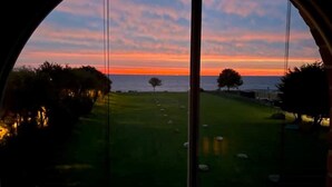 Une vue à travers une fenêtre circulaire sur un champ verdoyant, avec des arbres, et un coucher de soleil sur l’eau.