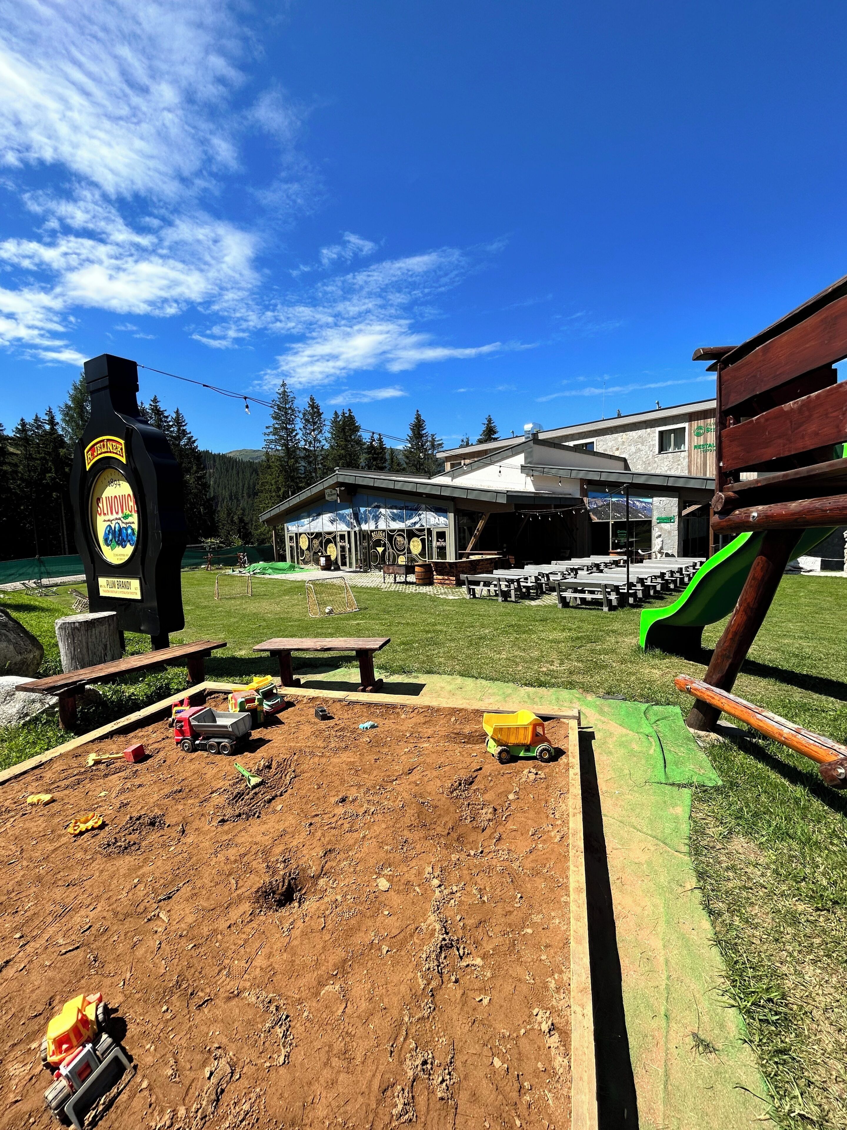 A dirt play area with toy vehicles, a wooden bench, and a picnic table. In the background, there's a building with a sign that reads "Restaurant."