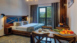 A bedroom with a wooden headboard, a bed with a blue and white bedspread, a dining table with fruit and cups, and a balcony with curtains. - HOTEL STRACHAN FAMILY JASNÁ (Demanovska Dolina)