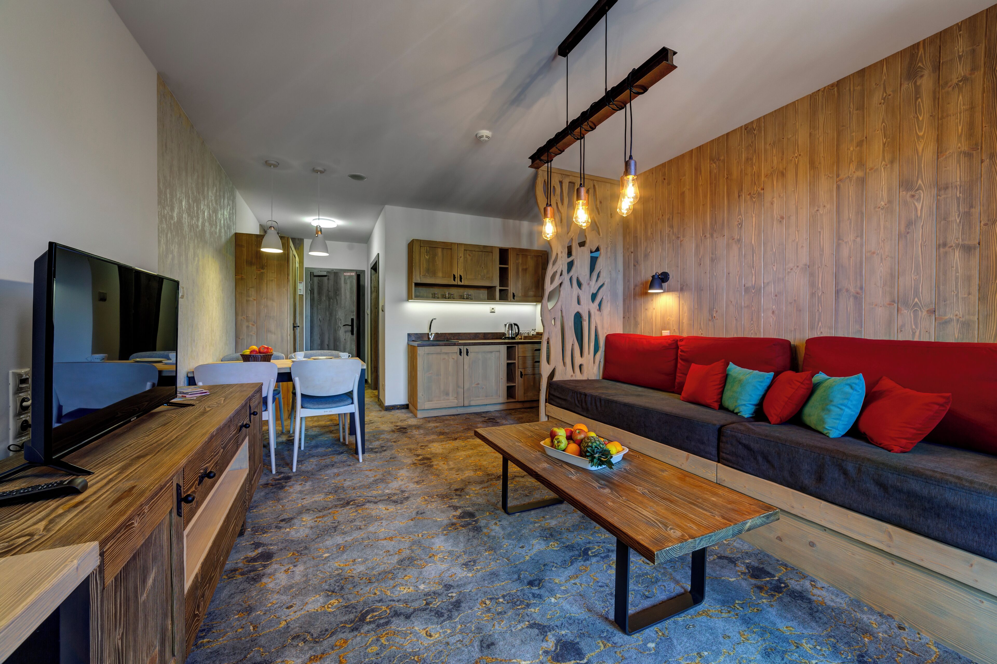 A modern living room with a wooden coffee table, a red sofa with colorful cushions, a television on a wooden cabinet, and a kitchen in the background.