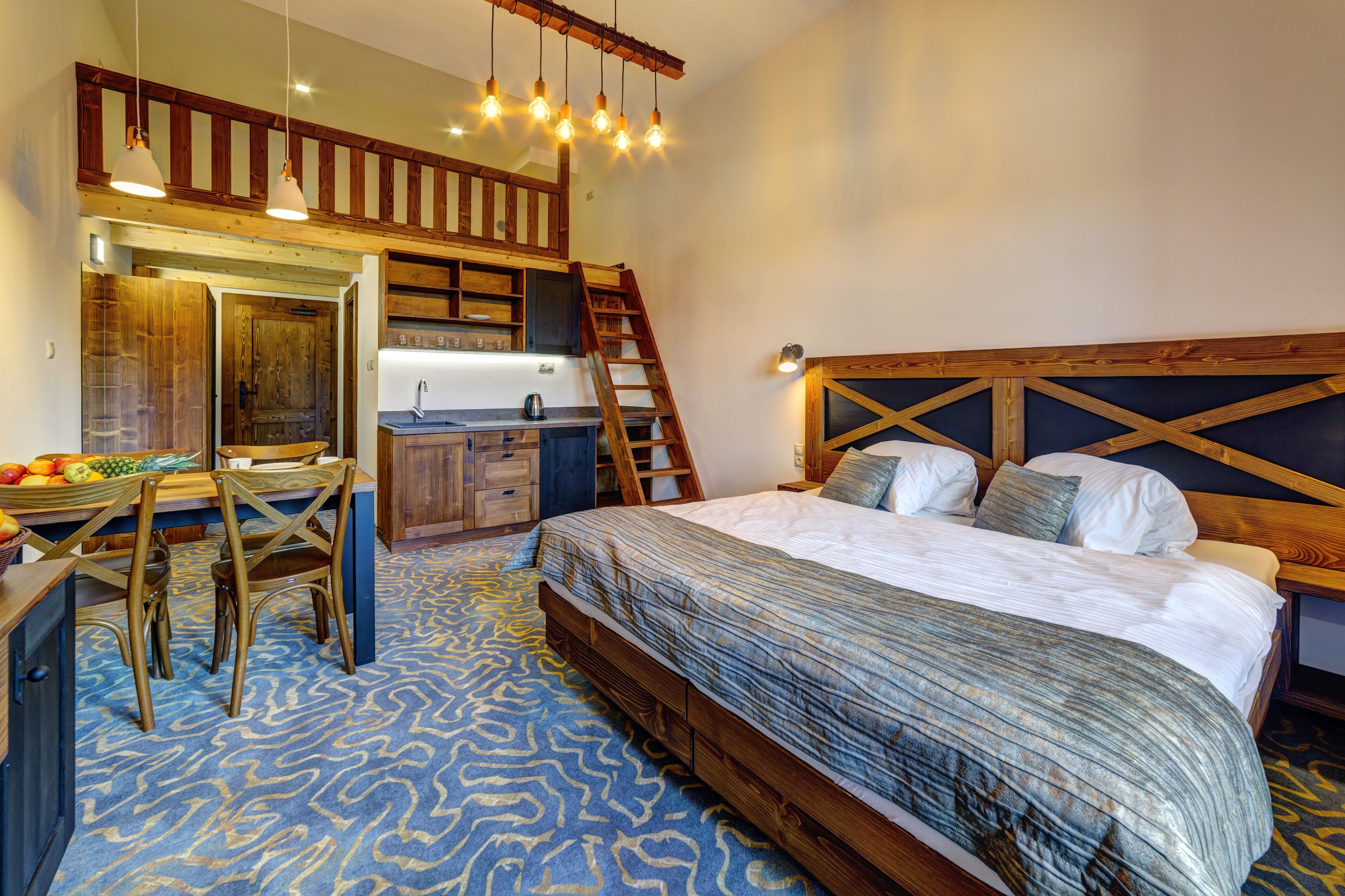 A bedroom with a wooden headboard, a bed with a patterned bedspread, a desk with a chair, a kitchen area with a wooden counter and cabinets, and a staircase.