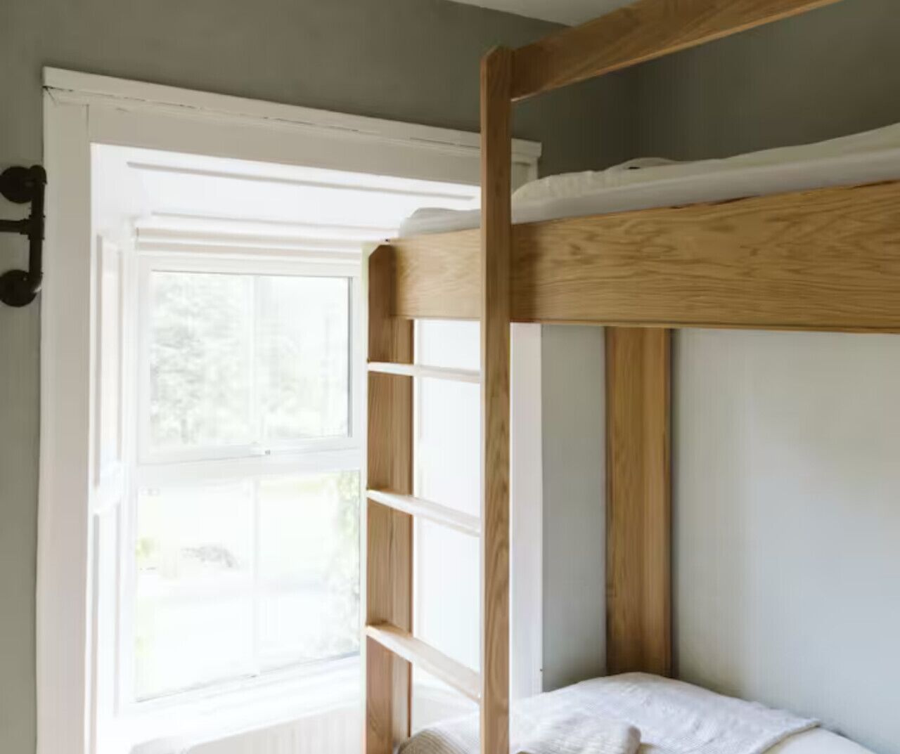 A bunk bed with a wooden frame and white bedding, a window with white blinds, and a white wall.