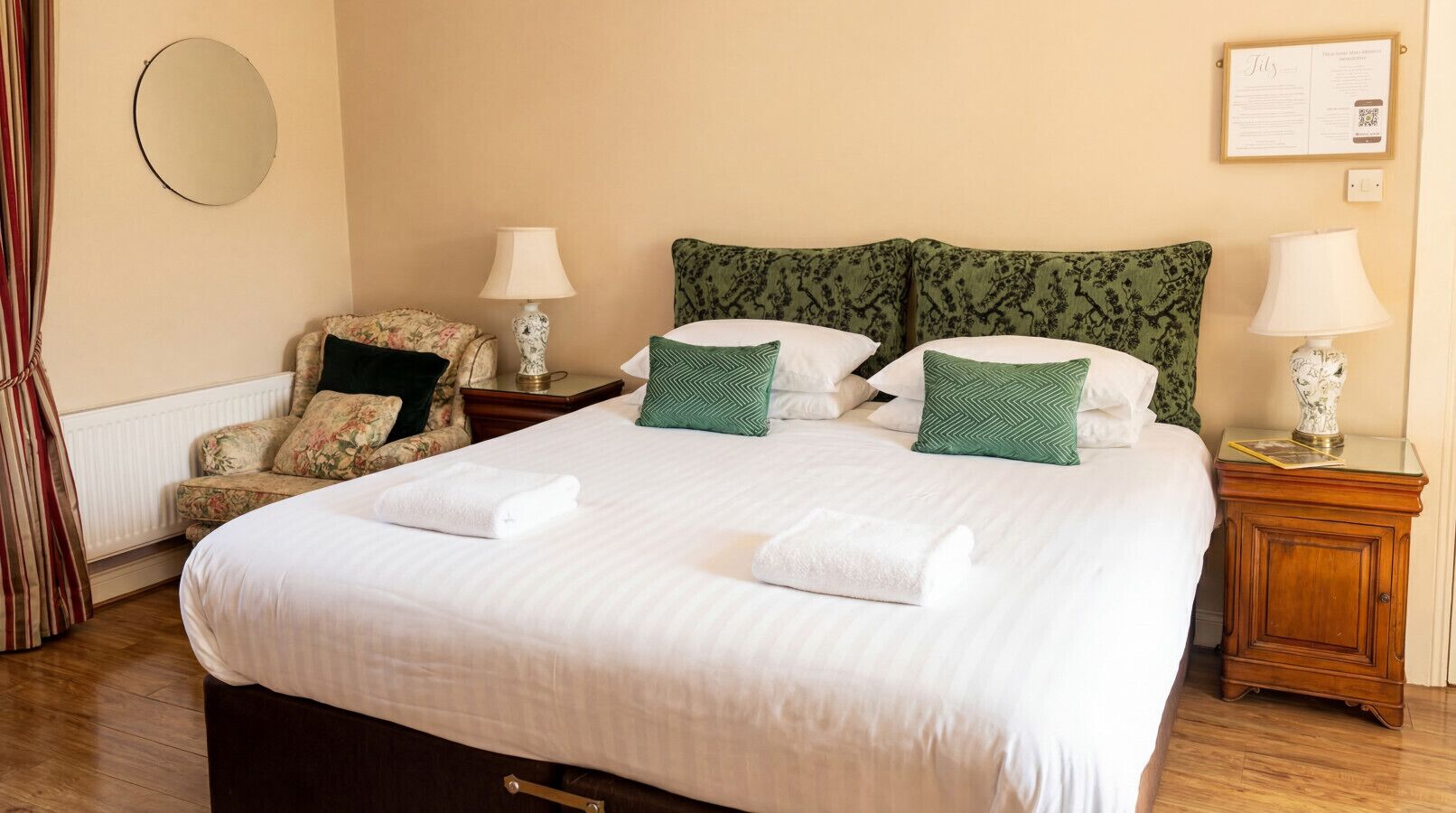 A neatly made bed with white linens, a floral armchair, a round mirror, and a framed certificate on the wall.