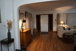 Een kamer met houten vloer, een witte deur, een bank, een lamp en een vaas met bloemen.